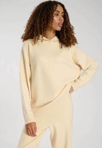 Sudadera crema hecha de una tela suave, con un corte holgado, hombros caídos y puños acanalados. Diseño minimalista con un cordón ajustable.