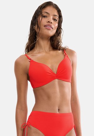 Frau mit nassen Haaren, die einen leuchtend roten Bikini mit verstellbaren Trägern und gerafften Seiten trägt, steht vor einem schlichten hellen Hintergrund.