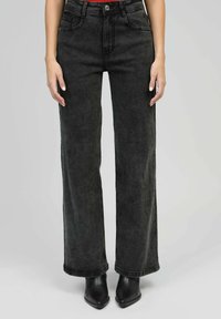 Femme portant un jean noir taille haute à jambes larges et des bottines noires à talons aiguilles, se tenant devant un fond simple.