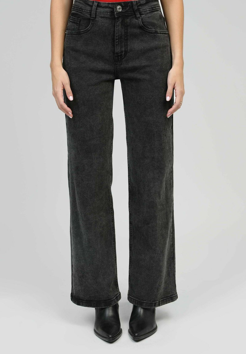 Femme portant un jean noir taille haute à jambes larges et des bottines noires à talons aiguilles, se tenant devant un fond simple.