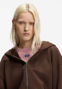 Bruine hoodie met ritssluiting en een gestructureerde afwerking, voorzien van een geborduurd logo en een brede capuchon. Draagt over een roze shirt met een blauw label.