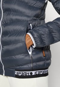 Dunkelgraue Pufferjacke mit horizontalem Steppmuster. Enthält weiße Akzente, Reißverschlusstaschen und ein schwarzes Band mit weißem Logo am Saum.