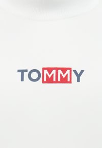 Bílá látka s výrazným vyšívaným logem "TOMMY" v modré a červené barvě, s červeným obdélníkovým rámečkem kolem "MM."