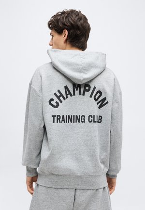 Young person wearing a gray hoodie and shorts with "Champion Training Club" printed on the back, facing away against a plain background.