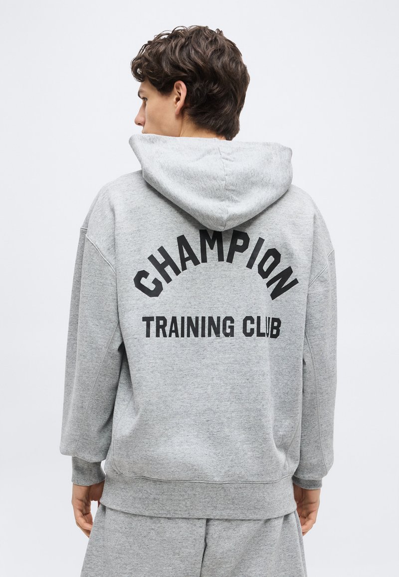 Tânăr purtând un hanorac gri cu glugă și pantaloni scurți, cu inscripția „Champion Training Club” pe spate, văzut din spate, pe un fundal simplu.