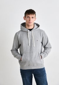 Felpa grigia in cotone con tasca frontale a marsupio, cappuccio con cordino e un piccolo logo sul petto. Abbinata a jeans blu.