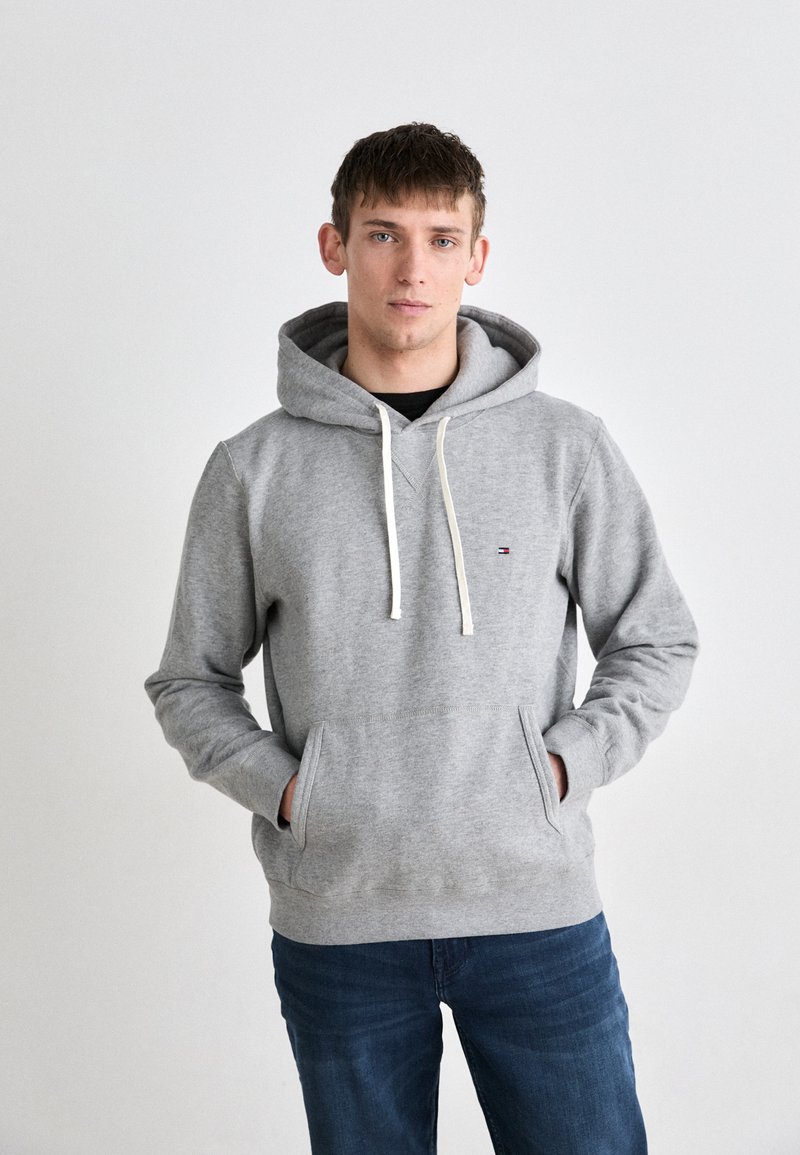 Felpa grigia in cotone con tasca frontale a marsupio, cappuccio con cordino e un piccolo logo sul petto. Abbinata a jeans blu.