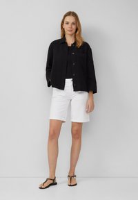Schwarze strukturierte Shirtjacke mit Kragen, Knopfverschluss und Tasche, kombiniert mit weißen knielangen Shorts und schwarzen flachen Sandalen.