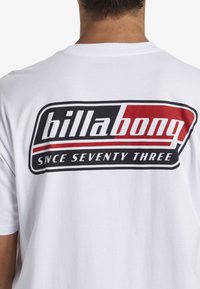 Bílý bavlněný tričko s velkým grafickým logem na zadní straně v černé a červené barvě, se slovy "billabong" a "SINCE SEVENTY THREE."
