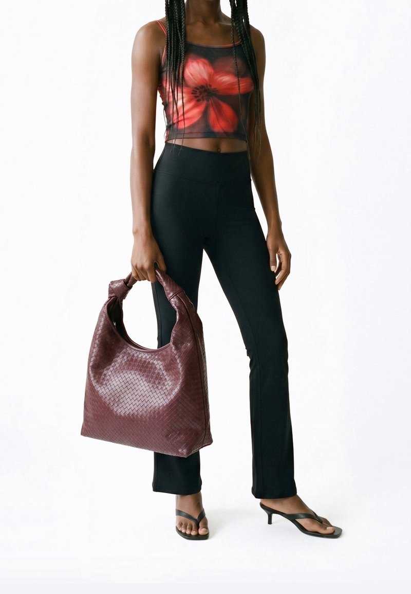 Personne portant un pantalon noir et un crop top rouge à motifs floraux, tenant un grand sac tressé rouge foncé et portant des sandales noires à talons.
