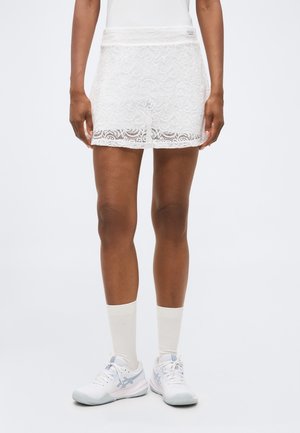 Femeie care poartă o fustă de tenis albă din dantelă, șosete albe până la gleznă și pantofi sport albi pe un fundal deschis simplu.