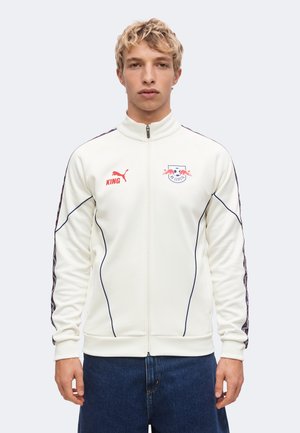 Young man wearing a white zip-up sports jacket with red Puma and RB Leipzig logos, standing against a plain light background.