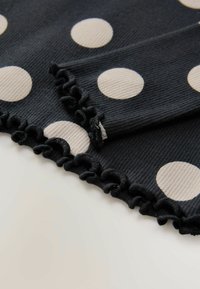 Mustaa kangasta, jossa on suuria valkoisia pilkkuja. Reunoilla on röyhelöitä kahdella sivulla. Materiaali näyttää olevan viirutettua ja siinä on tekstuurivaihteluita.