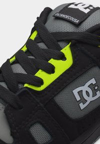 Κοντινό πλάνο γκρι και μαύρου αθλητικού παπουτσιού DC με φωτεινά κίτρινα οπές κορδονιών και μαύρα κορδόνια, που δείχνει το λογότυπο DC στη γλώσσα και στην πλευρική επιφάνεια.