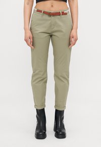 Chinos caqui com as bainhas dobradas, apresentando bolsos à frente e um cinto castanho com fivela. Botas pretas elegantes até o tornozelo completam o look.