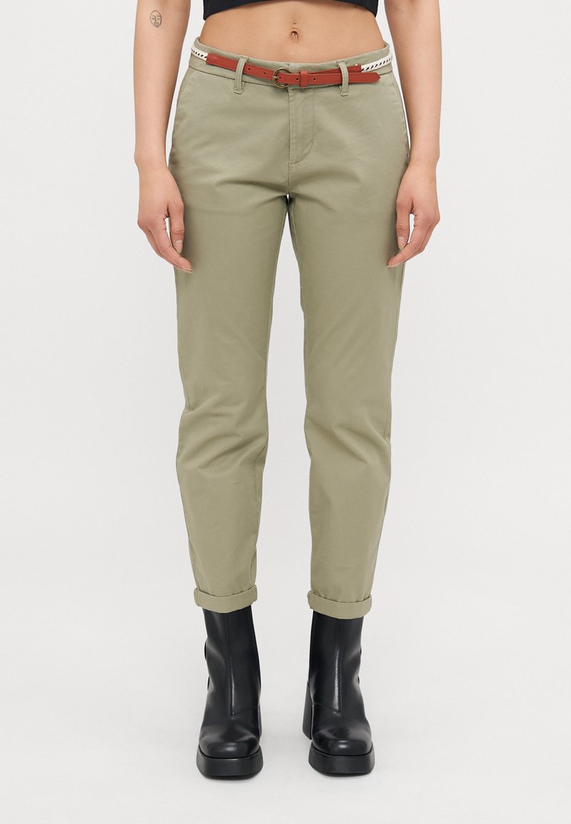 Chinos caqui com as bainhas dobradas, apresentando bolsos à frente e um cinto castanho com fivela. Botas pretas elegantes até o tornozelo completam o look.