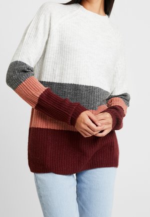 Femme portant un pull en maille côtelée avec des blocs de couleurs horizontaux en blanc, gris, rose et bordeaux, associé à un jean bleu clair.