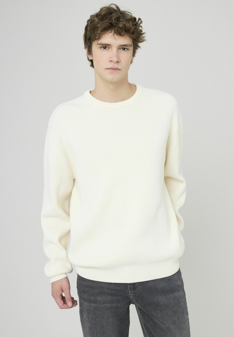 Pull en tricot blanc à côtes avec un col rond et des manches longues, associé à un jean gris foncé. Le tissu semble doux et texturé.