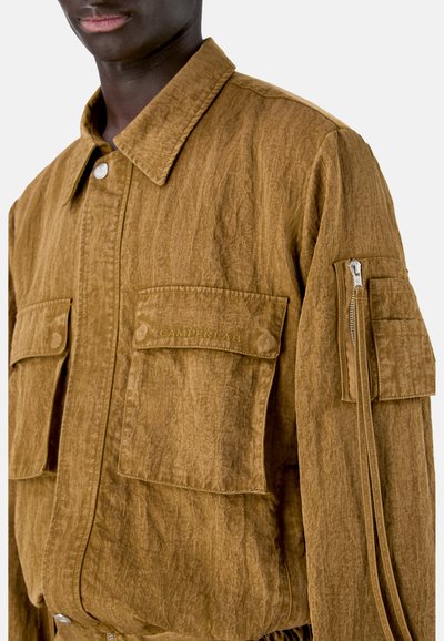 Primer plano de una chaqueta marrón con textura, con bolsillos de solapa en el pecho y la manga, llevada por una persona de piel oscura.