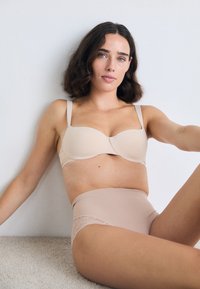 Γυναίκα με μαλλιά σε μήκος μέχρι τους ώμους και κυματιστά, καθισμένη απέναντι σε έναν λευκό τοίχο, φορώντας εσώρουχο σε nude απόχρωση και ψηλή εσώρουχα με δαντέλα.