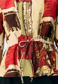 Multicolored blouse in lightweight fabric features abstract red, green, and beige patterns, with a cinched waist and decorative tie detail.