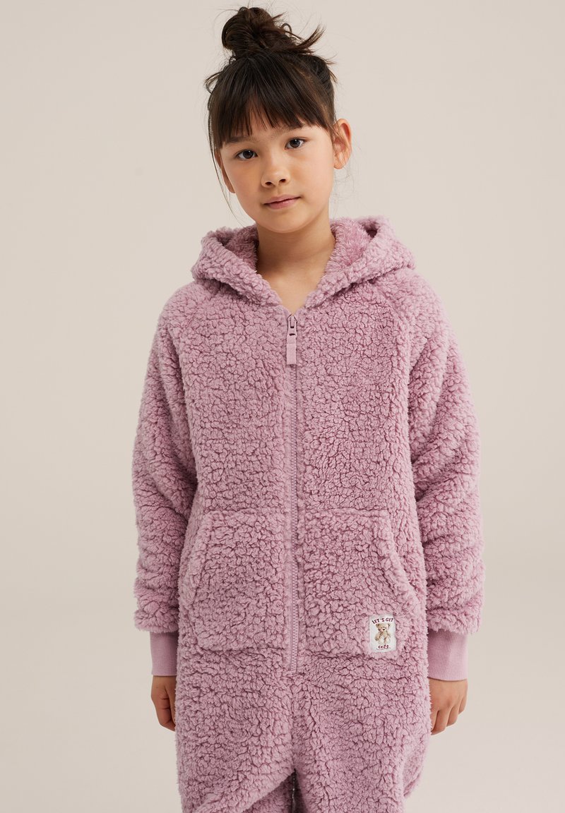 Jeune fille aux cheveux foncés coiffés en chignon, vêtue d'un sweat à capuche rose duveteux zippé avec poches avant et poignets côtelés, debout devant un fond uni.