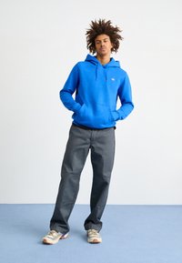 Sudadera azul con un bolsillo canguro, combinada con pantalones grises y zapatillas beige con acentos morados. Fondo simple, en pose de pie.