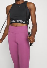 Personne portant un crop top noir Nike et des leggings violets, tenant des poignées de corde à sauter Nike noire et blanche.