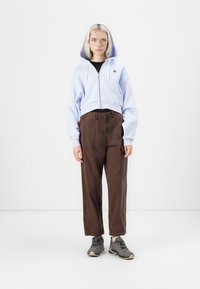 Sudadera recortada de color azul claro con cremallera y capucha, combinada con pantalones relajados de tiro alto en color marrón con cintura elástica y zapatillas grises.