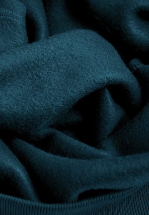 Teal fleece stof, zacht en textuurrijk, met zachte plooien. Zichtbare ribgebreide rand aan de onderkant. Rijke, diepe kleur zonder patronen.