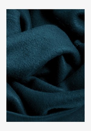 Teal fleece stof, zacht en textuurrijk, met zachte plooien. Zichtbare ribgebreide rand aan de onderkant. Rijke, diepe kleur zonder patronen.