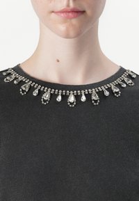Svart topp med en smykket kanten i krystaller med dråpe- og sirkelformede aksenter, flat tekstur og enkelt rund halsdesign.