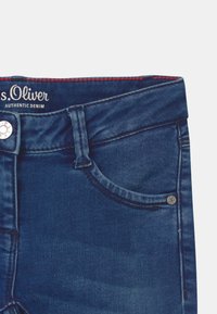 Jeans in denim blu scuro con una superficie liscia. Presentano un bottone in argento, cuciture sottili e una piccola tasca decorativa in metallo.