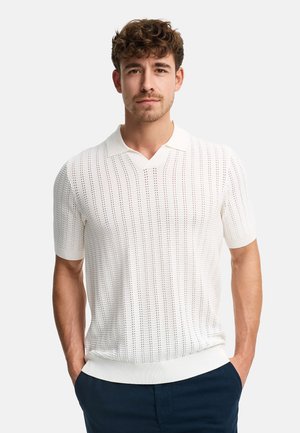 Man met kort krullend bruin haar, draagt een wit gebreid poloshirt met korte mouwen en donkere broek, staand met handen in de zakken.