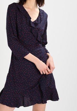 Femme portant une robe bleu marine à pois rouges, avec manches longues, un col en V volanté et un ourlet à volants.