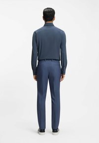 Man staat met zijn rug naar de camera, draagt een donkerblauw shirt met lange mouwen, blauwe op maat gemaakte broek en zwarte sneakers met witte zolen.