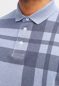 Polo en tissu bleu clair avec un motif à carreaux bleu foncé. Présente un col et trois boutons blancs. Design texturé.