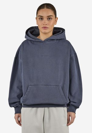 Mujer con una sudadera con capucha holgada de color gris oscuro con un bolsillo delantero y pantalones gris claro, de pie frente a un fondo liso.