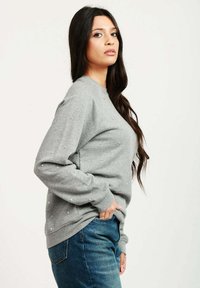 Felpa grigia con collo a giro, decorata con piccoli accenti simili a perle. Abbinata a jeans blu; mostra una vista laterale.