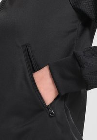 Svart idrottsjacka med slätt tyg, ribbade ärmslut och texturerade axeldetaljer. Har en blixtlåsficka på vänster sida.