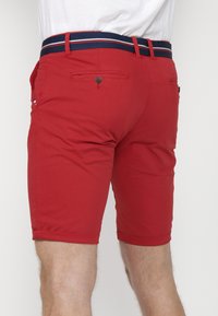 Shorts rouges en tissu léger, dotés d'une taille à rayures bleu marine et de poches. Les jambes sont ourlées juste au-dessus du genou.