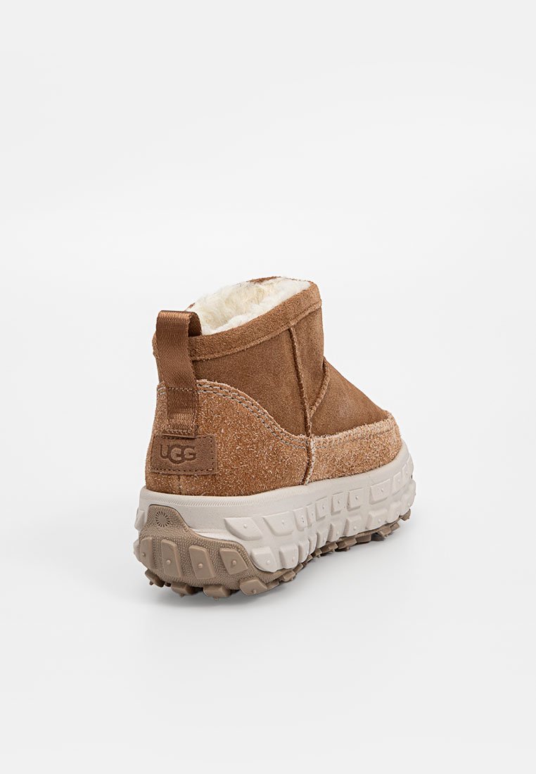 Brązowe zamszowe botki do kostki z fleece'owym wykończeniem i teksturowaną gumową podeszwą. Logo UGG widoczne z tyłu. Posiadają gładką cholewkę i zaokrąglony kształt.