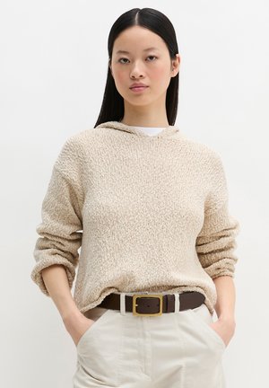 Femme aux cheveux noirs et lisses portant un sweat à capuche beige en tricot, un pantalon crème, et une ceinture marron foncé avec une boucle en or, les mains dans les poches.