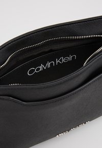 Černá kabelka na držení s texturovaným povrchem, zipovým uzávěrem a ozdobou ve stříbrném tónu na spodní straně. Uvnitř se nachází logo "Calvin Klein".