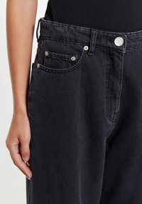 Lähikuva henkilön kädestä, joka lepää mustien, hopeisilla niiteillä ja napilla varustettujen farkkujen vieressä. Farkkuja on yhdistetty mustaan ribbineuleiseen toppiin.