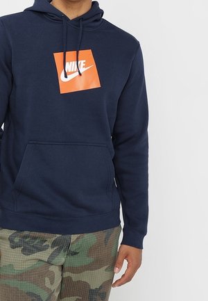 Osoba ubrana w granatową bluzę z kapturem Nike z pomarańczowym kwadratowym logo oraz spodnie moro, stojąca na białym tle.