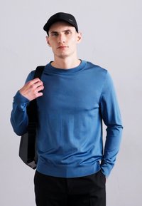 Mėlynas ilgomis rankovėmis marškinys su apvalia iškirpte, glotnia tekstūra ir raukšlėtu apvadu. Modelis laiko juodą rankinę prieš neutralų foną.