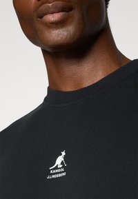 Melnas kokvilnas T-krekls ar apaļu apkakli, ar balto Kangol un J. Lindeberg logo apdruku uz krūtīm. Gluda tekstūra.