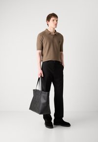 Brązowa koszulka polo z krótkim rękawem o fakturowanym wykończeniu, czarne spodnie eleganckie oraz duża czarna torba na ramię z wytłoczonym logo, noszona z czarnymi butami.