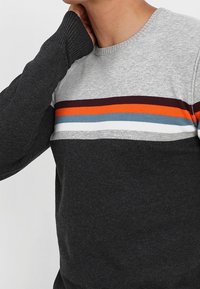 Pull gris foncé à col rond, avec des rayures horizontales orange, bleu, blanc et marron. Texture en tricot avec poignets côtelés.